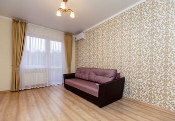An empty newly renovated room with light Wallpaper, curtains on the Oka and a laminate floor "under the tree". There is a brown sofa against the wall