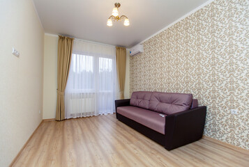 An empty newly renovated room with light Wallpaper, curtains on the Oka and a laminate floor "under the tree". There is a brown sofa against the wall