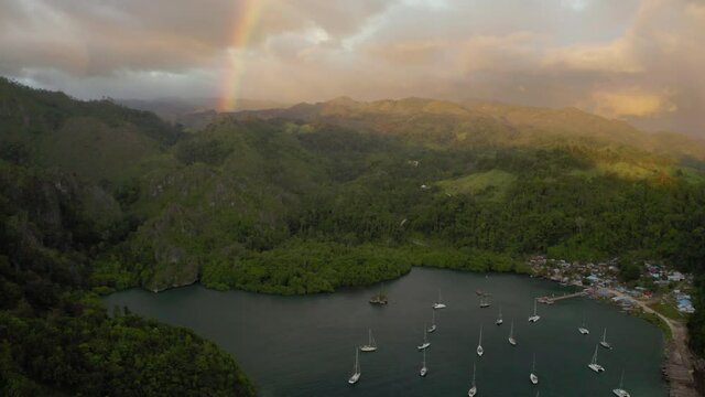 Aerial tilt up shot of harbor by village near rainbow against cloudy sky, drone flying over boats moored on sea near green mountains - Wakatobi Regency, Indonesia