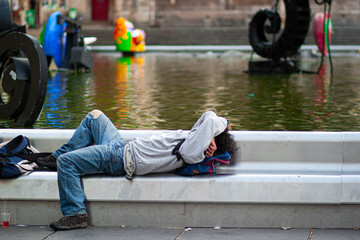 A homeless man wearing torn jeans is sleeping outdoor on a narrow metal bench by the river by covering his face with his arms. 