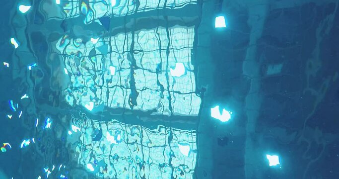 Under the water of an indoor swimming pool. Blue and aqua hues from the sloshing water are casted down from the sunlit roofs.