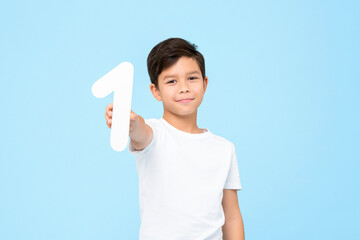 Portrait of smiling young Asian boy holding and showing number one cutout in isolated studio blue...