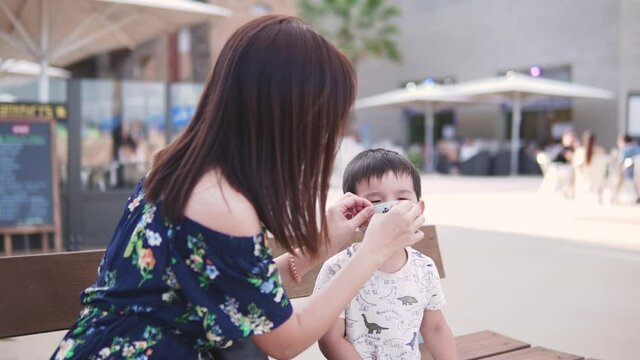 Young mother putting face mask on her son face. Sitting outdoors wihile preventing contamination. New normality after coronavirus pandemic in Spain.