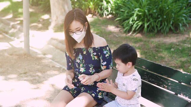 Young Asian woman wearing face mask and spraying hand sanitizer alcohol on her son hands at the outdoor park. Protection and preventive measures new normality after coronavirus pandemic