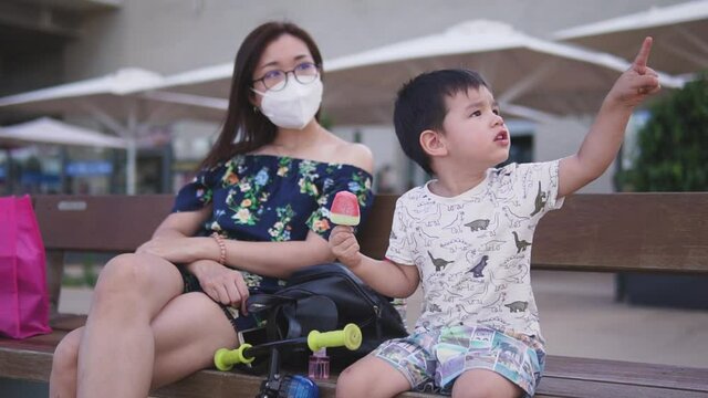 Mother and son enjoying a day outdoors wearing face mask. New normality video. Life after coronavirus. Eating ice cream on a bench at the beach. Social distancing.