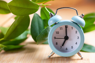 Alarm clock morning wake-up time. Blue alarm clock on table with green leaves on background....