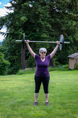 Middle aged caucasian woman with gray hair lifting a barbell with black plates, fitness outside on the lawn
