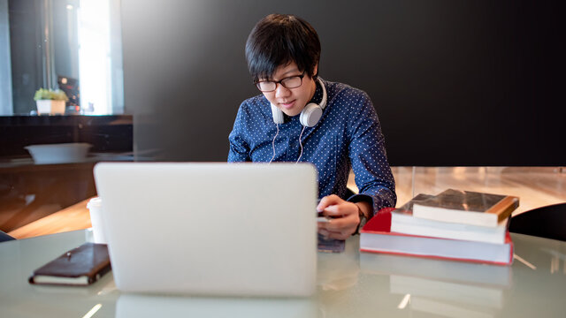 Smart Asian business man with glasses and headphones using laptop computer while reading book in public library. Male freelance working from coworking space. Internet of things concept. - Powered by Adobe