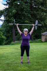 Middle aged caucasian woman with gray hair lifting a barbell with black plates, fitness outside on the lawn
