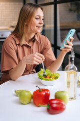 Young woman using her mobile phone while eating salad in cozy wooden kitchen. Concept of healthy eating.