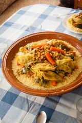 Vertical shot of table served couscous tagine on a blue squared tablecloth. Dish ingredients chickpeas, carrot, pumpkin, cauliflower, turnip, beef meat and semolina cooked on a big size ceramic plate.