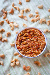 Top view shot of a small blue and white bowl with vegan sobrasada or cashew dip on a green striped tablecloth with dry tomato, paprika, olive oil, garlic and pepper and surrounded by  dry cashews.