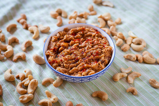 Food plated shot of a small blue and white bowl with vegan sobrasada or cashew dip on a green striped tablecloth with dry tomato, paprika, olive oil, garlic and pepper and surrounded by  dry cashews.