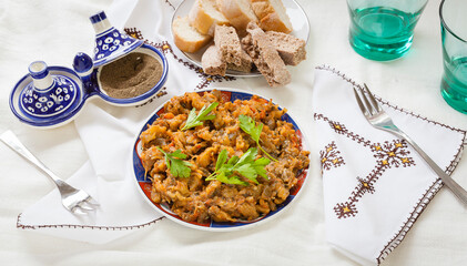 Top view food shot of typical Moroccan mashed aubergine and tomato with spices salad Zaalouk plate with parsley leaves and homemade rye bread plated on a traditional tablecloth linens with forks
