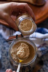 Vertical shot of a spoon filling a crystal pot with ginger powder spice on a home traditional kitchen by unfocused woman hands. Exotic and authentic flavors stills with negative space.