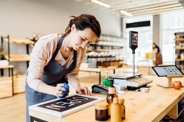 Lovely woman owner of zero waste shop in grey apron preparing , writing open sign on tablet sign in front of counterbar.social distancing concept,coronavirus is outbreak in city.