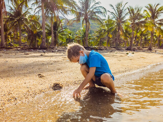 Cute little young blond boy with medical face mask on the beach playing with sand. Protecting from air pollution and virus infection as new normal lifestyle during the covid19 virus epidemic era