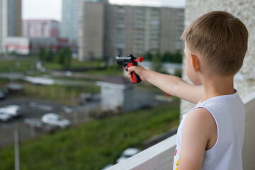 A small boy shoots a toy gun, standing in the loggia of a multi-storey building. The manifestation of aggression in children.