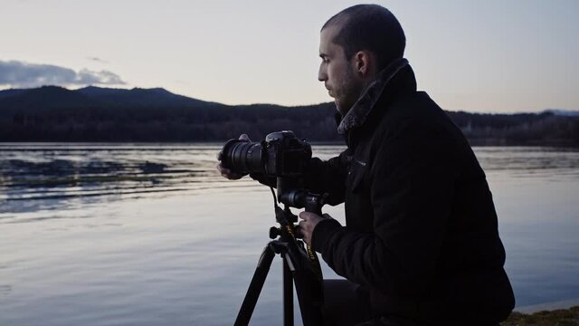 Caucasian man adds filter to camera lens attached to tripod by calm lake waters with dark mountain range in background, static profile close up