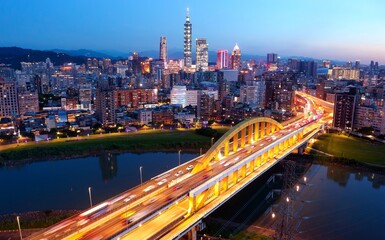 Night skyline of Taipei City viewed from above a riverside park with a highway bridge spanning Keelung River &  towers standing out amid the skyscrapers in Xinyi Commercial District under sunset sky