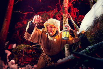 Obraz premium Evil medieval monk in canvas sackcloth robe with lattern in dark forest with snow and red light on winter night. Fantasy or fairy tale about wandering monk. Forces of good and evil in world