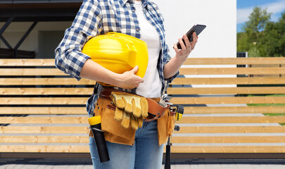 repair, construction and building concept - woman or builder with smartphone, helmet and working tools on belt over living house background
