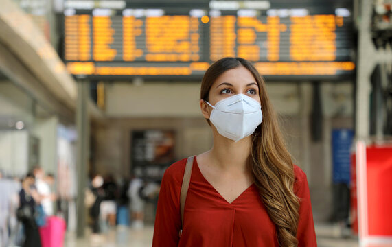 Traveler woman wearing KN95 FFP2 face mask at train station to protect from virus and smog. Young caucasian woman with timetables information of departures arrivals on behind.
