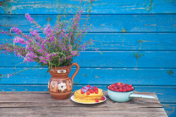 Crepes with fresh raspberry, old bowl and bouquet with field flowers on wooden table. Blue wall on the background.