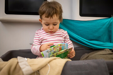 little cute toddler sitting in the baby bassinet of an airplane sitting and reading a children's...