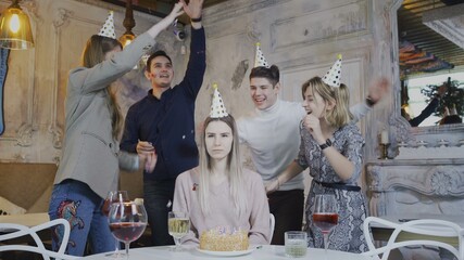 Sad birthday girl is sitting at the table while friends congratulate her