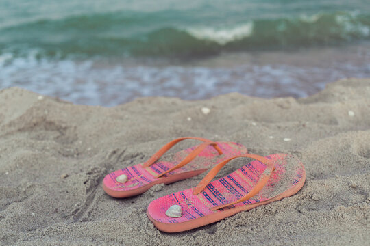 Left pink orange ornamental female shoes flip flops at sandy beach with sea waves view and shells