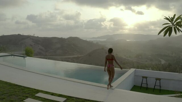 Aerial shot of woman walking by infinity pool during sunset, drone flying forward towards sea and mountains against sky - Lombok, Indonesia