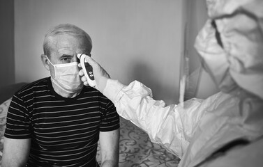 Black and white snapshot of a doctor's examination at patient's home during quarantine 2020. Cropped image of doctor's hands measuring old man's fever with infrared thermometer. Coronavirus concept
