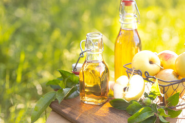 Two glass bottles of apple cider vinegar and fresh ripe apples on a wooden nature background. Copy...