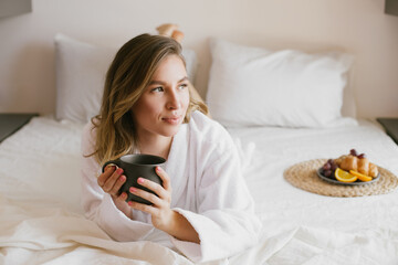 Young beautiful woman wearing white bathrobe having breakfast in bed with coffee and croissant and fresh fruits in cozy bedroom.