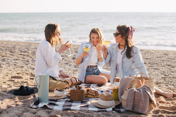 Three young beautiful happy women female friends having cozy summer picnic with lemonade, fresh bread and fruits on a beach.