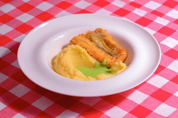 Fried fish with mashed potatoes and sauce served on a white plate over red plaid tablecloth.