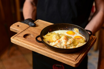 Close up shot of waiter serving a plate of traditional breakfast with fried eggs in a pan on a rustic wooden board.