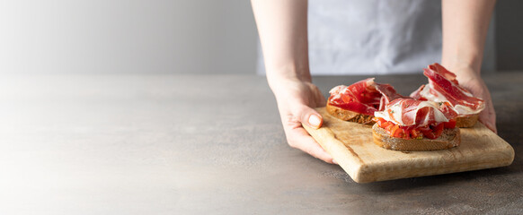 Woman holding wooden try with toasted brad slice with fresh tomatoes and cured ham. Italian...