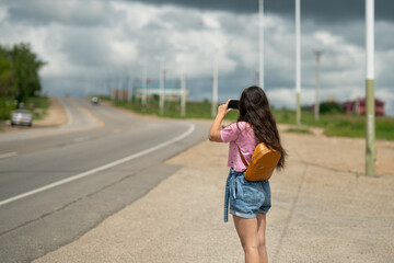A girl with long hair in a pink T-shirt stands near the road  and takes pictures on the phone