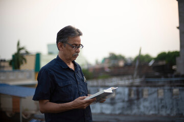 An old / aged Indian Bengali man in blue shirt is reading a book while standing on a rooftop under the open sky. Indian lifestyle and seniors