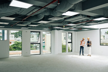 Horizontal extreme long shot of two modern architects standing together in unfinished room discussing building plan, copy space