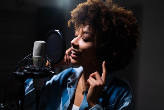 Close up of an young professional smiling energetic african female singer wearing headphones is performing a new song with a microphone while recording it in a music studio. 