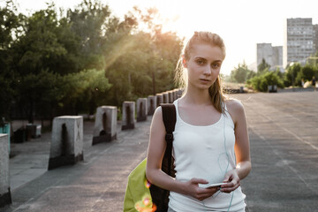 Beauty young female model with long blond hairstyle in sport clothes and with backpack standing on the road and looking sideways. The girl listen to music in earphones and walking on the cort