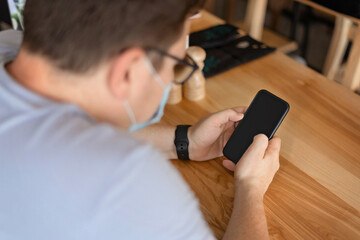 Caucasian man sitting in restaurant and using phone in medical face mask. New Normal concept
