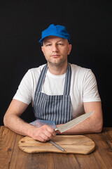 Fototapeta premium Portrait of professional butcher in white t shirt, black and white apron and blue baseball hat sitting at table and holding knife in his right hand, small cutting board in front of him.
