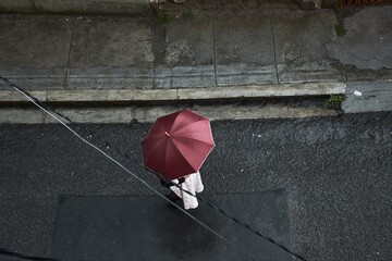 High angle shot of a woman holding a dark red umbrella and walking in the street on a rainy day © Miguel Ángel Mamani Valdemira/Wirestock