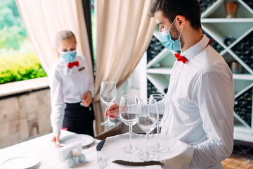 A waiter in a medical protective mask serves a table in the restaurant. Employees of a restaurant or hotel in protective masks. The End Of Quarantine.