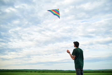 Jun 5, 2020-Ternopil/Ukraine:Young brunette skinny man, wearing dark green t-shirt, playing with colorful kite on green field meadow in summer. Kite flying in blue cloudy sky. Family leisure activity