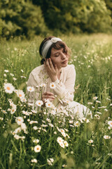 Young beautiful woman in white clothes and white headband with decoration on summer nature in the park with daisies.
